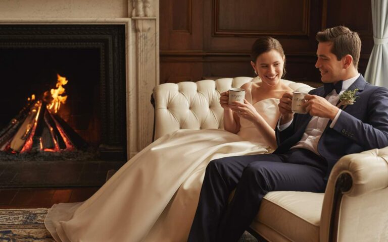 A photograph of a smiling couple, both around 32 years old, relaxing on a plush, cream-colored velvet couch at the end of their wedding day. The bride is wearing a flowing ivory gown while the groom is in a dark navy suit, both holding steaming mugs of coffee and sharing a quiet moment of contentment. Behind them, a warm, crackling fire burns softly in a grand Australian heritage-style fireplace, casting a gentle glow on the room which features dark wood paneling and antique furnishings. Soft, diffused light from a nearby window creates a cozy and intimate atmosphere. less